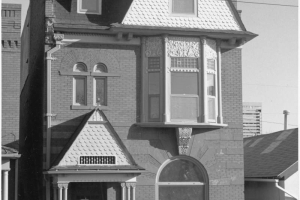 View of a three story brick house at 2418 Stout Street in the Five Points neighborhood of Denver, Colorado. The house has eclectic architectural elements that include arched windows with voussoirs, bargeboard gables, corbiestep sides, plaster relief details, and a dormer. A sign in the yard reads: "Offered by Marsh & Associates, 628-5474, For Rent."
