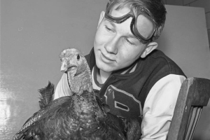A teenage Red Cross volunteer poses with a live turkey, probably in Denver, Colorado. The boy wears a team jacket emblazoned with the letter "B." He has a pair of goggles on his head.