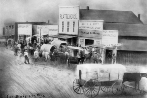 Painting of the corner of Blake and 15th (Fifteenth) streets, in Denver, Colorado. Shows covered wagons, people, and false front businesses. Signs read: "Meat Market, Shaw-Bailey & Co.," "Keith & Bond, Auction, Commission & Storage," "Daniels & Brown," "Platte House," "Ford's Barbershop," "Heatley Chase," "[?] Beer," and "Empire Bakery."