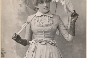 Studio portrait of Mrs. Leonel Ross Anthony O'Brien holding an umbrella with lace trim. She wears leather gloves and a dress with a pleated waist line, buttons, and lace collar.