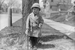 A boy poses outdoors with a cane possibly in Denver, Colorado. He wears a coat and bowler.