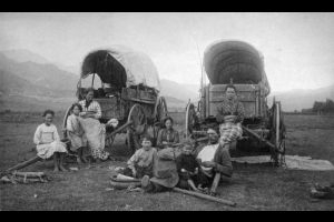 Weary pioneers rest in front of two Conestoga wagons on a plain with mountains in the background. Family includes an adult male, 3 adult females, and 4 children.