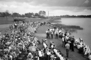 Young boys with toy sailboats gather at the shore of Sloans Lake at Sloans Lake Park in the Sloan Lake neighborhood of Denver, Colorado. The school band is seated with instruments in hand. Students and observers crowd near the shore. Three African-American (Black) students are in the crowd.