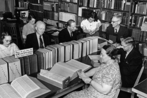 Men and women work in the Bibliographic Center in the Denver Public Library at Colfax Avenue and Bannock Street in the Civic Center neighborhood of Denver, Colorado. Books and stacks are nearby. A sign reads: "Please Do Not Close the C.B.I. and U.S. Catalogs Turn Pages Carefully."