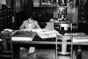 A man reads at a table in the Western History department of the Denver Public Library at Colfax Avenue and Bannock Street in the Civic Center neighborhood of Denver, Colorado. Maps and large, bound books are on the table. Bookcases, filing cabinets, and model of a Colorado Midland locomotive and coal car are in the room.