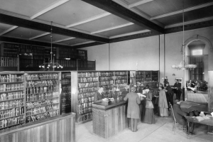 Interior view of the Denver Public Library at East Denver High School in Denver, Colorado; shows people at a counter, bookshelves, a coffered ceiling and hanging lamps. John Cotton Dana is behind the desk.