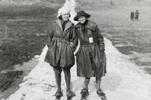 Women wearing skis pose on a snow path at Genesee Park (Jefferson County) Colorado; outfits include leather winter coats and a knit hat. Grass meadow is to the sides.