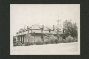 View of the Sisters of Loretto Academy and convent in Las Vegas (San Miguel County), New Mexico. Shows an adobe building with siding and Territorial style architectural elements. The building has a portal, a balcony, dormers, chimneys, and a wooden steeple topped with a cross. A woman poses on the balcony. Trees and bushes are in the yard.