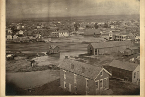 View of flooded Cherry Creek and inundated buildings in Denver, Colorado, Shows brick and frame houses and commercial buildings. Signs read "American House," "Commonwealth," and "J.G. Vawter & Co." The South Platte River and the Front Range are in the distance.