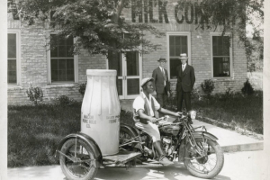An African American man rides an Indian Motorcycle with a Winter-Weiss Company platform sidecar in front of the Macon Pure Milk Company in Macon, Georgia. He wears a cap, vest, gaiters add boots. An advertising model of an oversized milk bottle is on the sidecar's platform. Lettering on the bottle reads: "Macon Pure Milk Co." Two men in suits and hats look on.