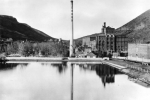 View east of the Adolph Coors Company Brewery, between Table Mountain, Golden, Jefferson County, shows lake in front of the multi-story buildings, tall "Coors" smokestack and wooden fence.