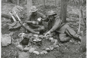 Two men relax near a camp fire, and cook bacon, in a forest probably on Mount Bierstadt in Clear Creek County, Colorado. The men wear outdoor clothing, high leather boots, and hats. One man pours condensed milk into a metal drinking cup. Metal camping utensils, canvas bags and an axe are near the camp fire ring.