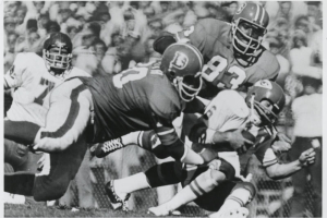 Two Denver Broncos professional football players tackle an opponent. Spectators are in the background.