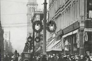 Pedestrians walk along bustling Sixteenth Street around noon at the intersection of Stout Street in the City and County of Denver, Colorado.  Christmas wreaths hang from the lightposts.  Cars and bicycles are parked along the street.  Landmarks include the Daniels and Fisher Tower.