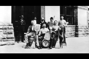 Group of men pose outside the Adolph Coors Company brewery, Golden, Colorado; they hold long handled whisk brooms, mugs of beer, and shovels with "Wet Golden 1909," "Malt House 1909" and "Section 11 Brewery," lettered on them. Lennard Vogel sits on a wooden barrel keg on the left, Alphonse Thuet stands in the back right.
