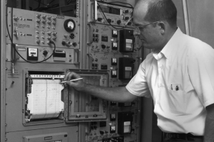At the National Bureau of Standards in Boulder, Colorado, John Milton targets a rolling graph paper with a mechanical pencil. The date on the paper is: "Jul 23 1968." The glass, metal, and rubber gasket cover is opened, and the surrounding metal frames have electronic components mounted in them. Gauges, toggle switches, dials, digital (mechanical) readouts, and RF cables front each of the instruments.
