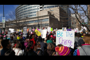 Large group of protesters by the Wellington Webb Building. Sign in the foreground "Be the Change"