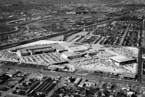The zig-zag floor plan of Cinderella City shopping center, opened in 1968, is visible in an aerial view. The huge complex of connected two- story buildings and parking in Englewood, Colorado, is between Santa Fe Drive and Elati Street, and Hampden Avenue and Floyd Ave.; residential neighborhoods surround the area. Signs on buildings include: Cinema," "Woolworth's," "Western Federal Savings," "Englewood."
