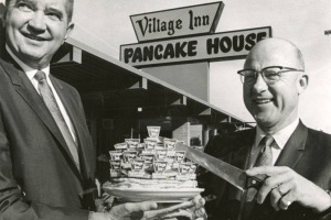 Merton S. (Andy) Anderson, left, vice-president, and Jim P. Mola, president of the Village Inn pancake house restaurants, celebrate the 10th birthday of their franchise operation with a birthday cake made of pancakes. The restaurant behind them at 8855 E. Colfax Ave., was the first in the group which now numbers 37 stores.