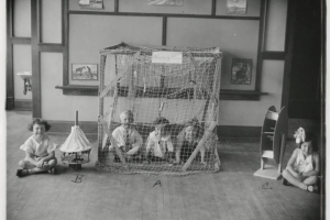 Photograph of five Barnum Elementary Students in Denver, Colorado.  Three of the students are in a "Monkey Cage", or a wood frame with a string net, one girl is next to a miniature carousel and another is next to a Ferris wheel made out of wood and cardboard.  Below the three objects are the letters "A", "B", "C".