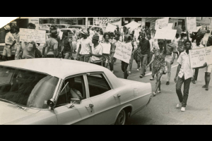 From back of photograph: Demonstrators march against traffic during trek Saturday to Police Bldg. Lt. Jerry R. Caroccia drives police car.