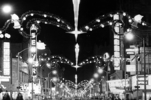 Nighttime view of 16th (Sixteenth) Street in downtown Denver, Colorado. Automobiles drive under Christmas lights and decorations that span the street. Signs read: "Denver," "Baurs," "Douglas Optical," and "Paramount."