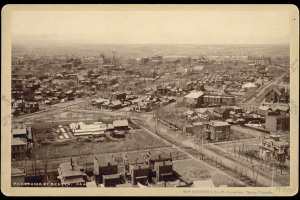 Aerial view of 19th (Nineteenth) and Welton Streets, frame and brick houses, and commercial buildings in downtown Denver, Colorado. Buildings in the distance include: Union Station, the Tabor Block with a square corner tower, the Windsor Hotel, Arapahoe School, and the stone First Presbyterian Church.