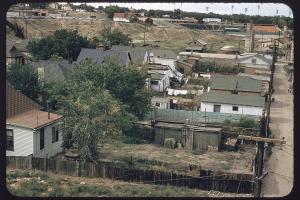 View of Bear's Stadium in the Sun Valley neighborhood, Denver, Colorado. The stadium is one level built into a hillside on the site of a landfill. Shows the backyards of houses, clothes lines, an abandoned building with smoke stacks and houses on Federal Boulevard.