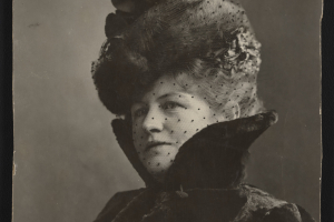 Leonel O'Bryan, otherwise known as Polly Pry, sits for a studio portrait wearing the late Victorian or Edwardian style clothing. Her outfit features a large hat adorned with silk flowers or faux foliage and a high collared coat. A sheer, black veil with chenille dots covers her face.