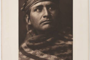 Portrait of a Navajo (Dine) man. He wears a cloth headband and a woven patterned blanket around his shoulders. Titled by Curtis: A chief of the desert - Navaho