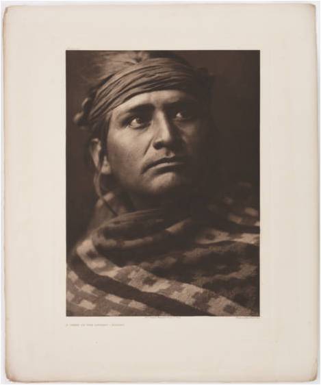 Portrait of a Navajo (Dine) man. He wears a cloth headband and a woven patterned blanket around his shoulders. Titled by Curtis: A chief of the desert - Navaho